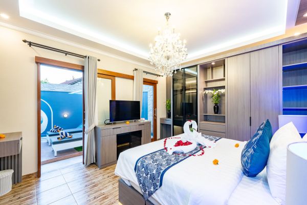 Luxurious Balinese villa bedroom with king bed adorned in rose petals and towel swan, open doors to blue-tiled pool terrace, chandelier, and built-in wardrobes.